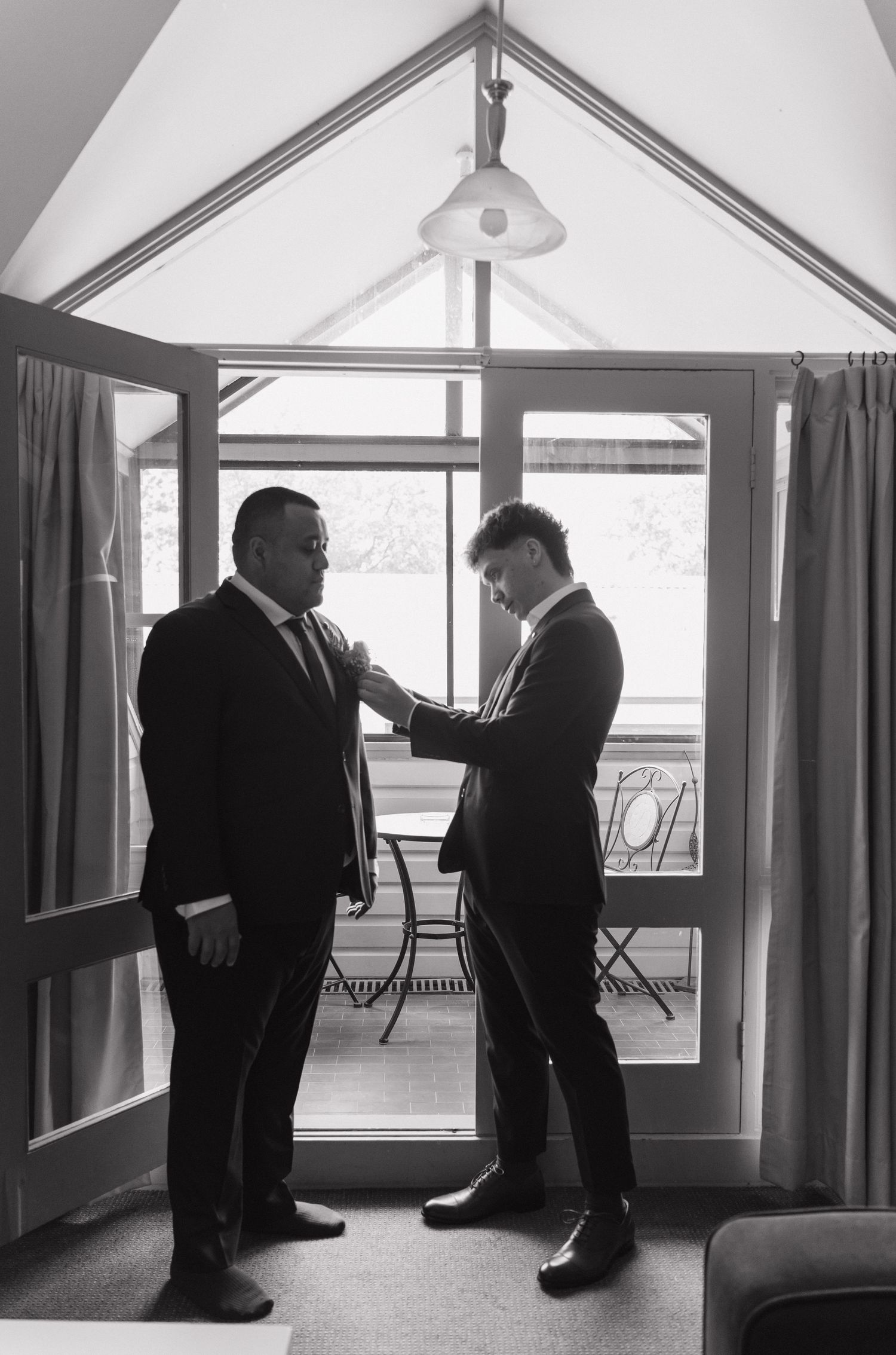 Two men in suits helping each other prepare in a sunlit room, captured in black and white wedding photography.