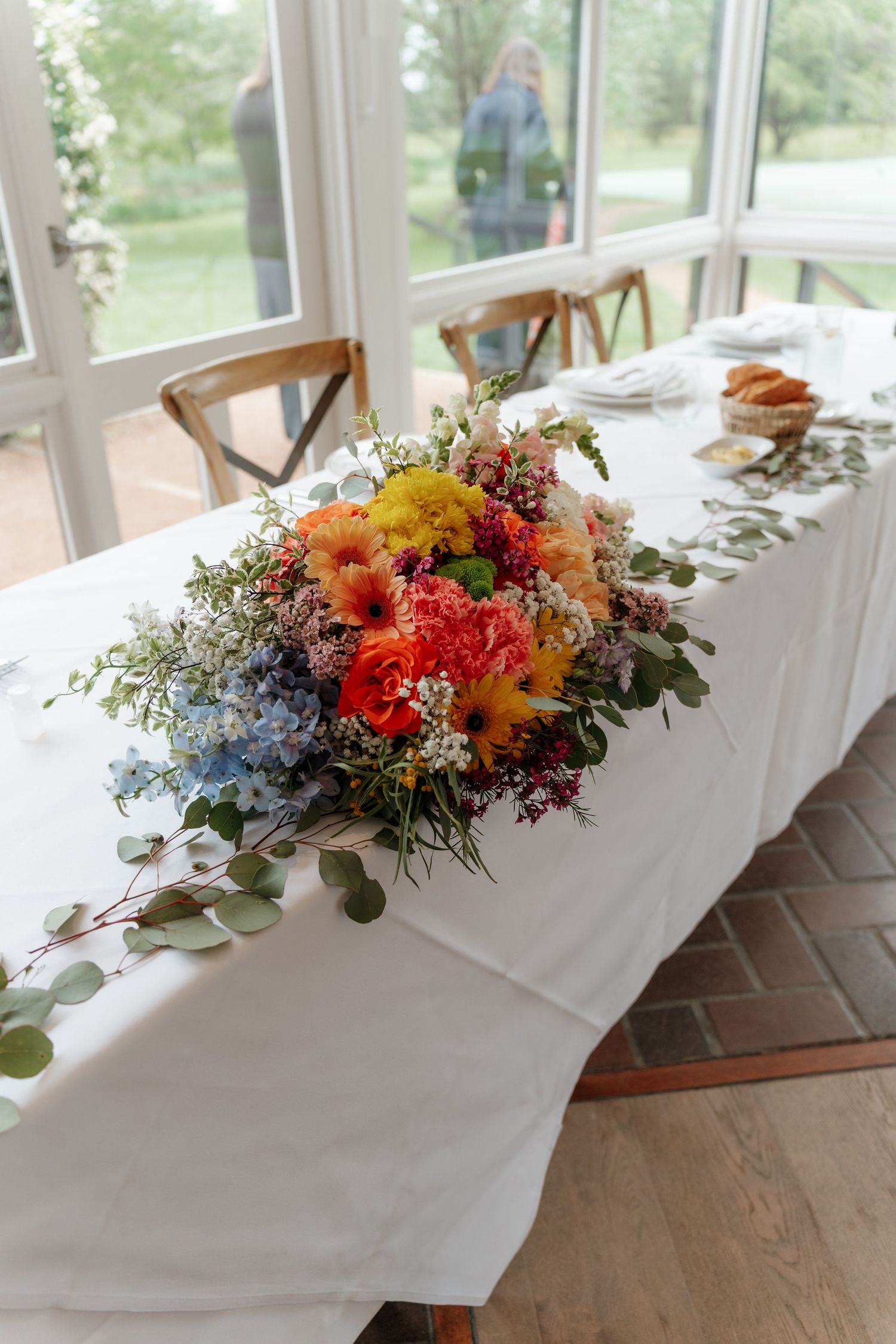 Colorful floral centerpiece with orange, yellow, and blue blooms on a white tablecloth at an elegant event.