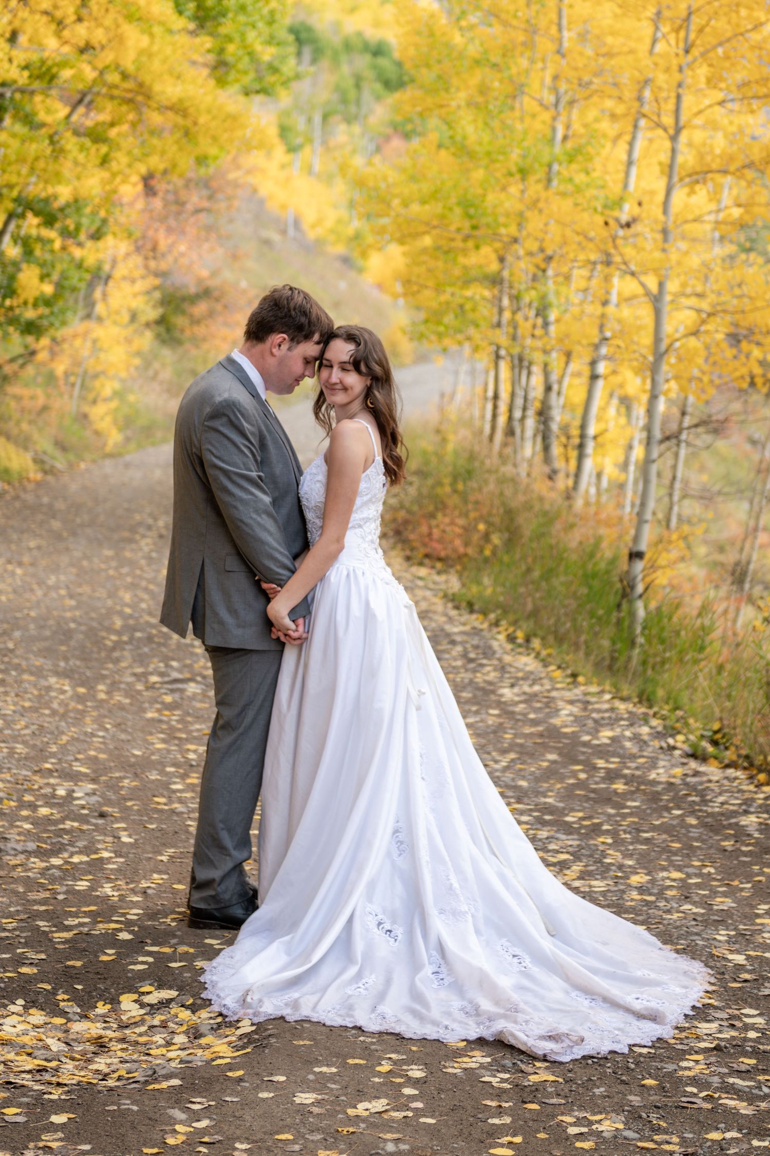Bride and groom embrace on a leaf-covered path surrounded by golden autumn aspen trees in a romantic fall wedding photo.