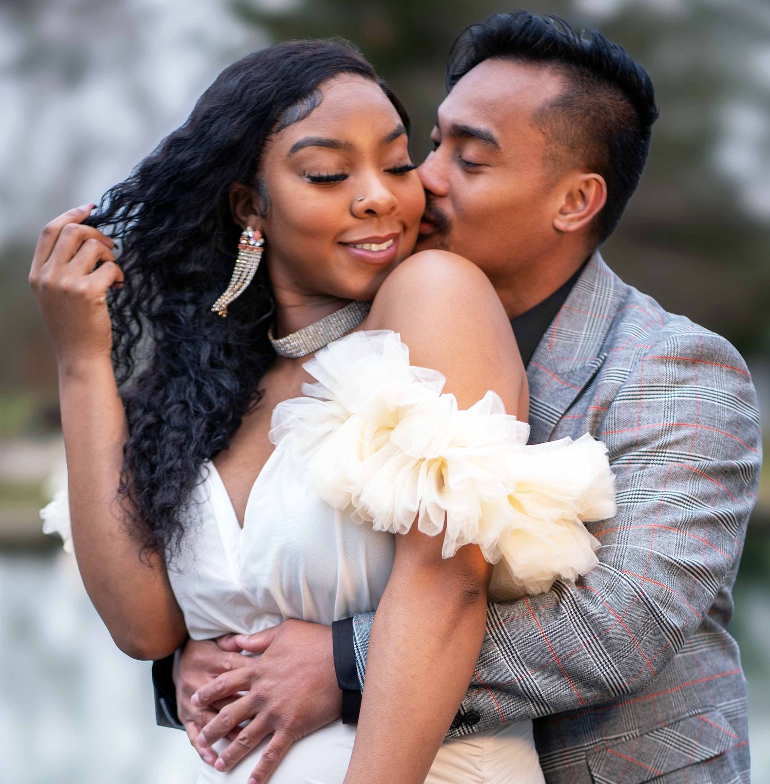 A couple shares a romantic embrace outdoors, man kissing woman's cheek, both dressed elegantly for an engagement photo session.