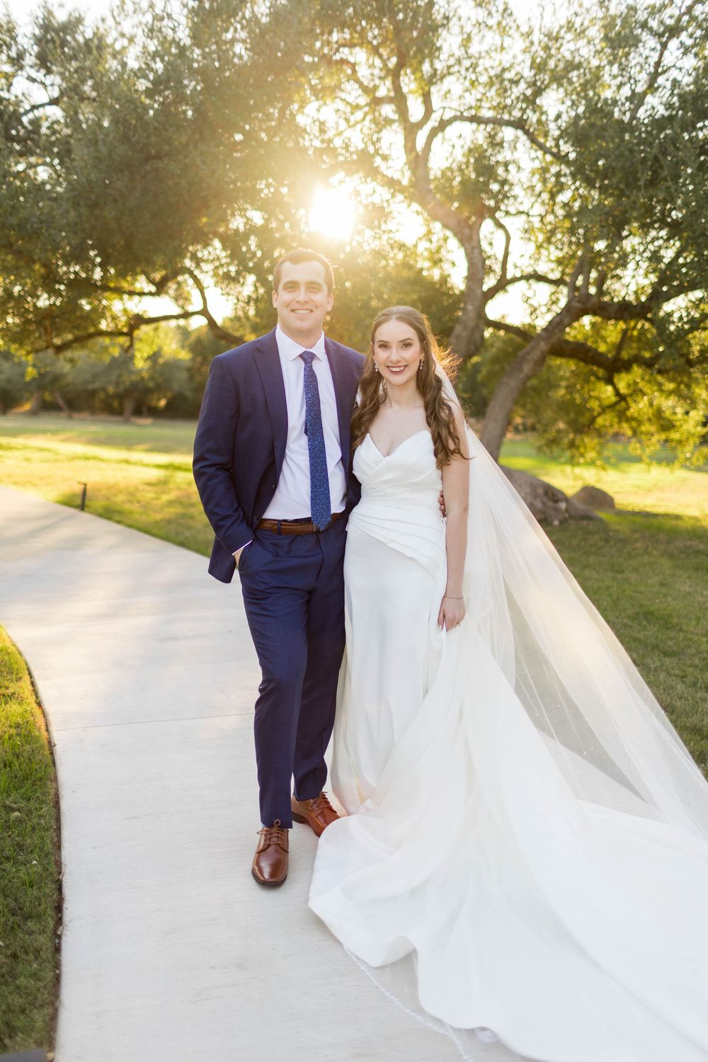 Bride and groom pose on a sunlit path at golden hour, bride in white gown with veil, groom in navy suit.