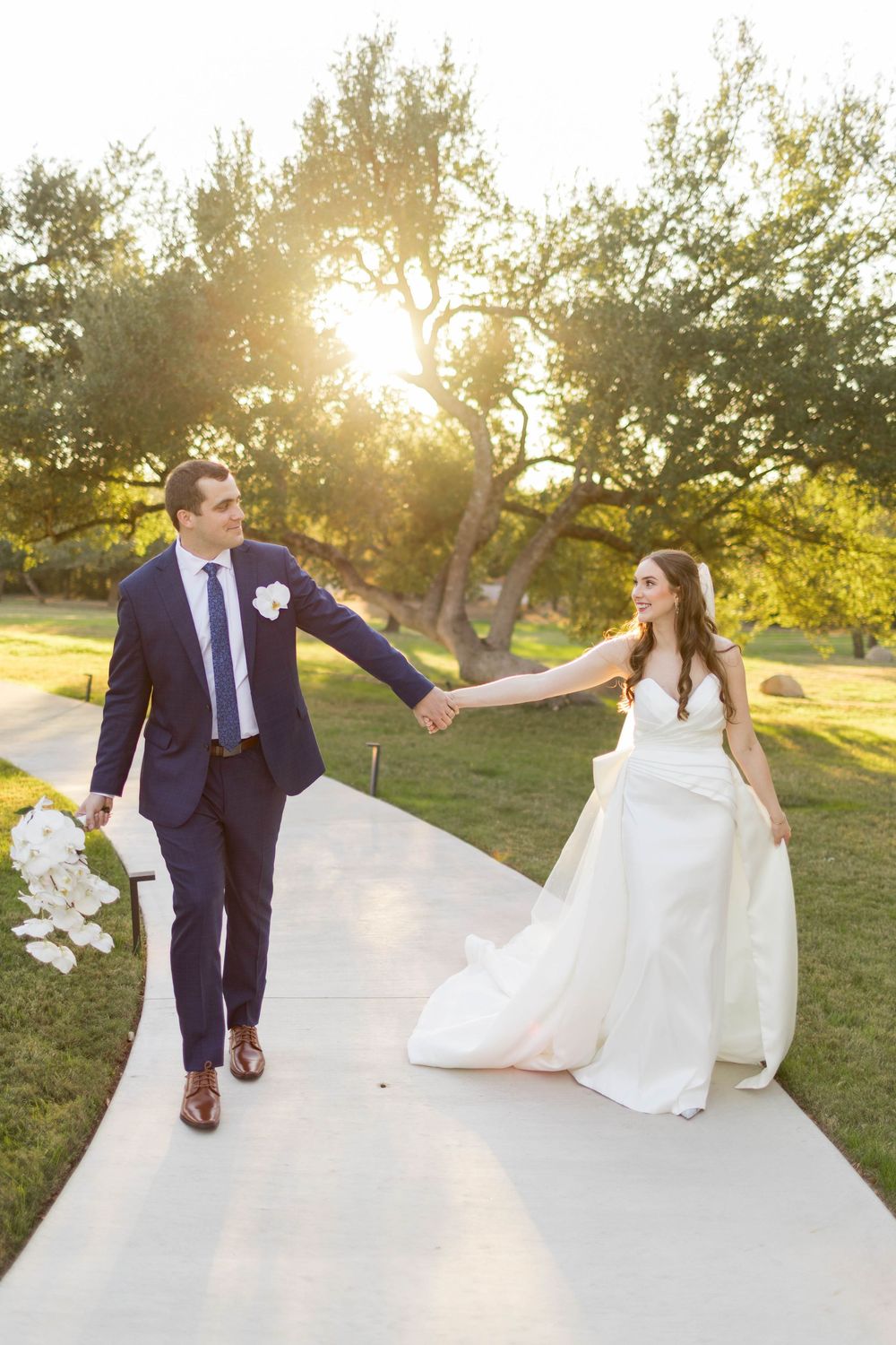 Bride and groom hold hands walking down a sunlit path at golden hour, surrounded by lush green trees.