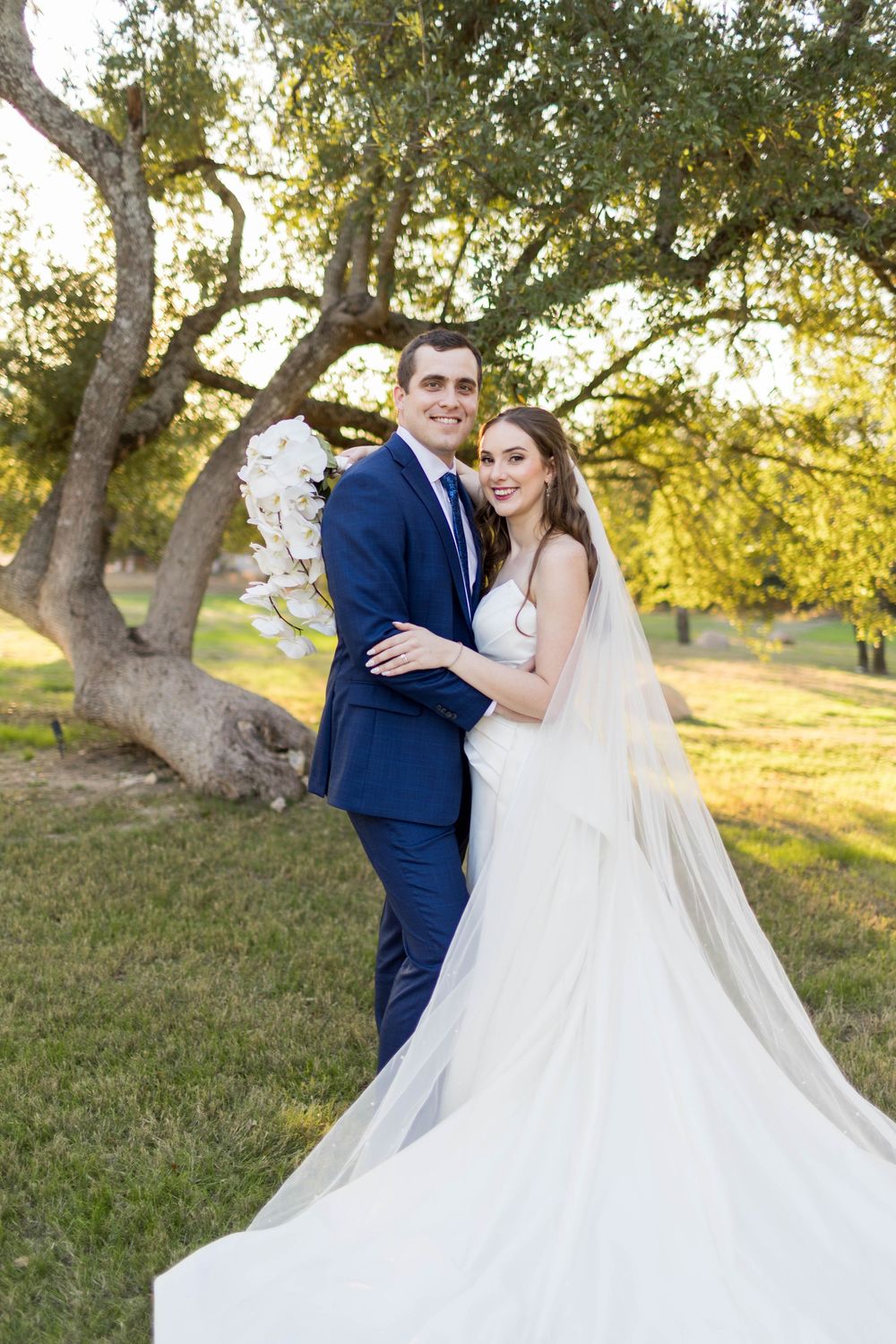 Bride in white gown and veil poses with groom in navy suit outdoors under oak trees on their wedding day.