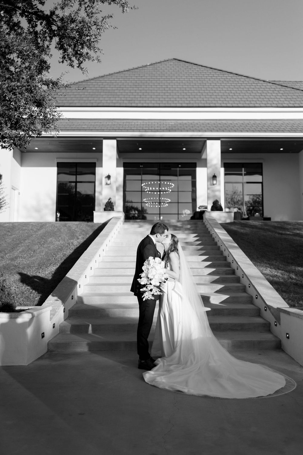 Black and white photo of bride and groom kissing on grand staircase steps outside elegant wedding venue.