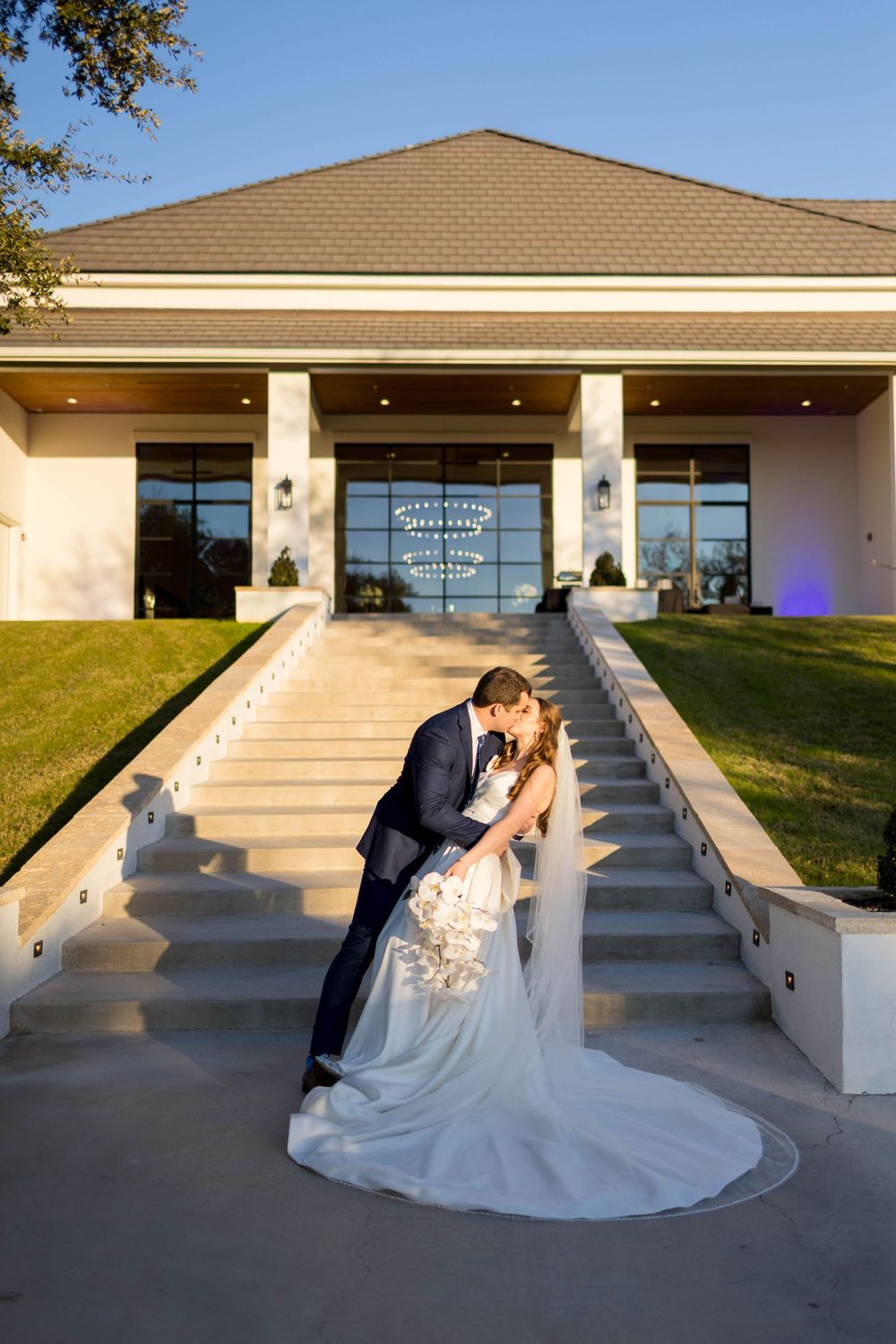 Bride and groom share a romantic kiss on grand outdoor staircase of elegant wedding venue at golden hour.