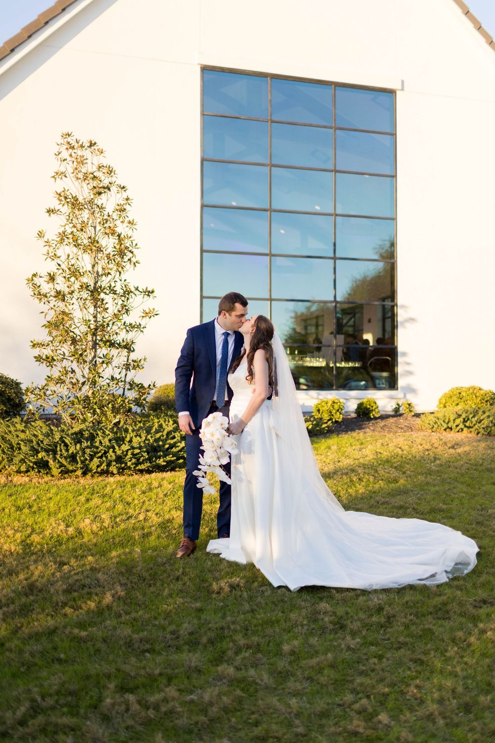 Bride and groom share a tender kiss on green lawn outside a modern white wedding venue building.