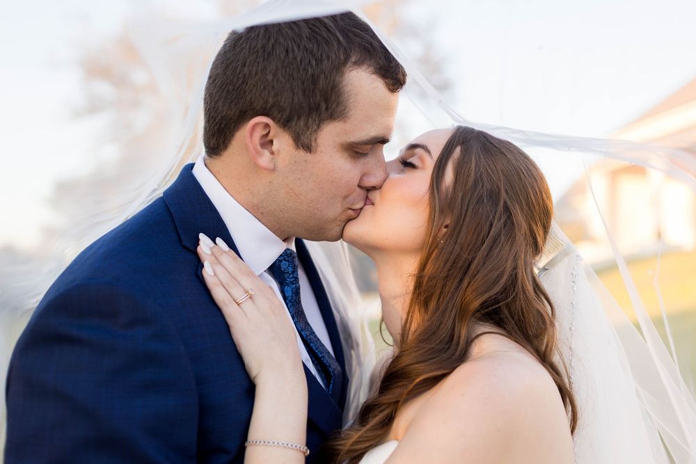 Bride and groom sharing a romantic kiss under a white veil, groom in navy suit, bride in white wedding dress.
