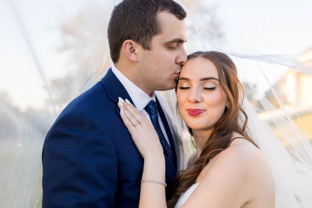Groom in navy suit kissing bride on cheek, bride smiling with eyes closed in romantic outdoor wedding portrait.