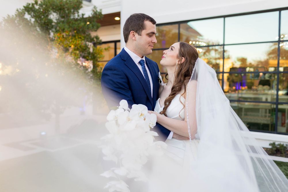 Bride and groom share a romantic moment outdoors, with her white veil flowing in the foreground at their wedding.