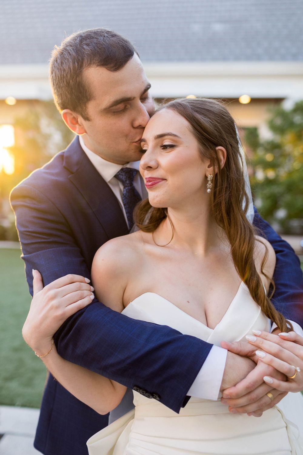 Groom in navy suit kisses bride on temple at golden hour, embracing her in white strapless wedding gown.