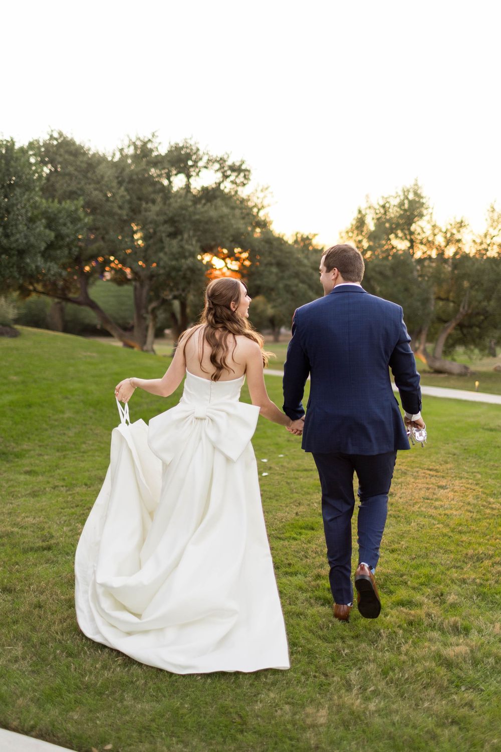 Bride and groom walking hand in hand across green lawn at sunset, bride in white gown with bow, groom in navy suit.