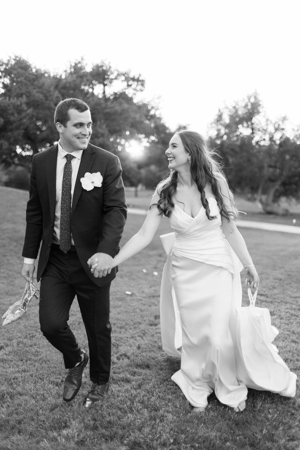 Bride and groom walk hand-in-hand across green lawn in black and white wedding portrait at sunset.