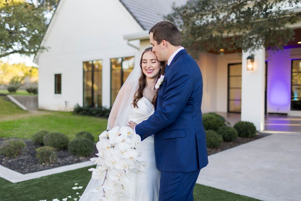 Bride and groom share a romantic embrace outside a white farmhouse venue, bride holding a lush white floral bouquet.
