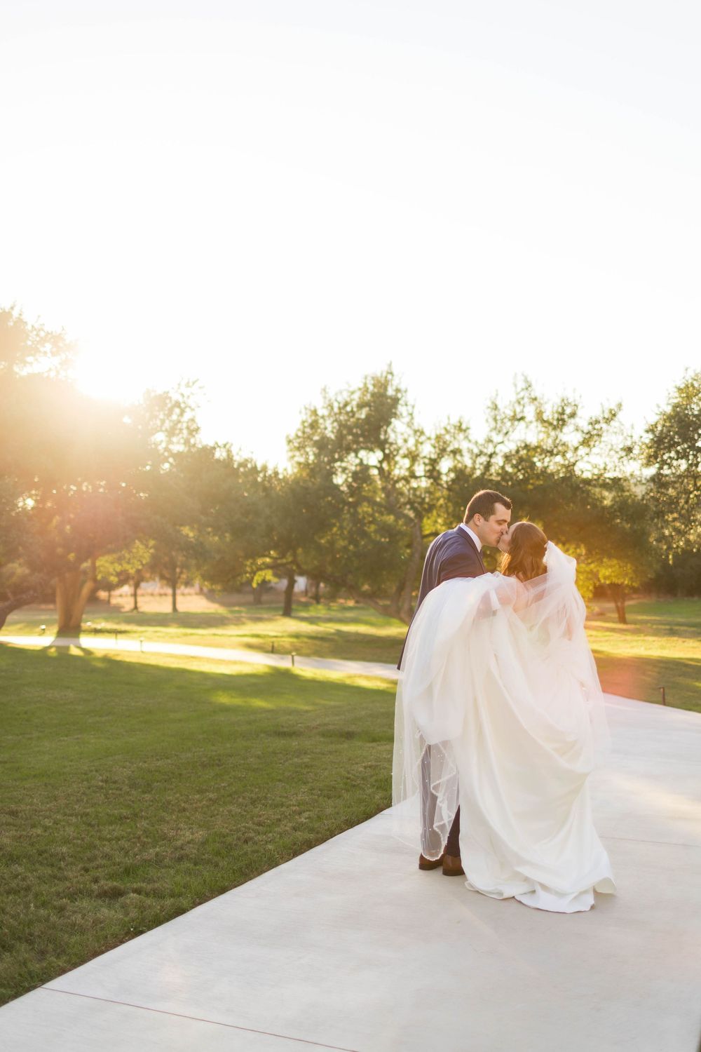 Bride and groom share a romantic kiss at golden hour on a sunlit path surrounded by lush green trees.