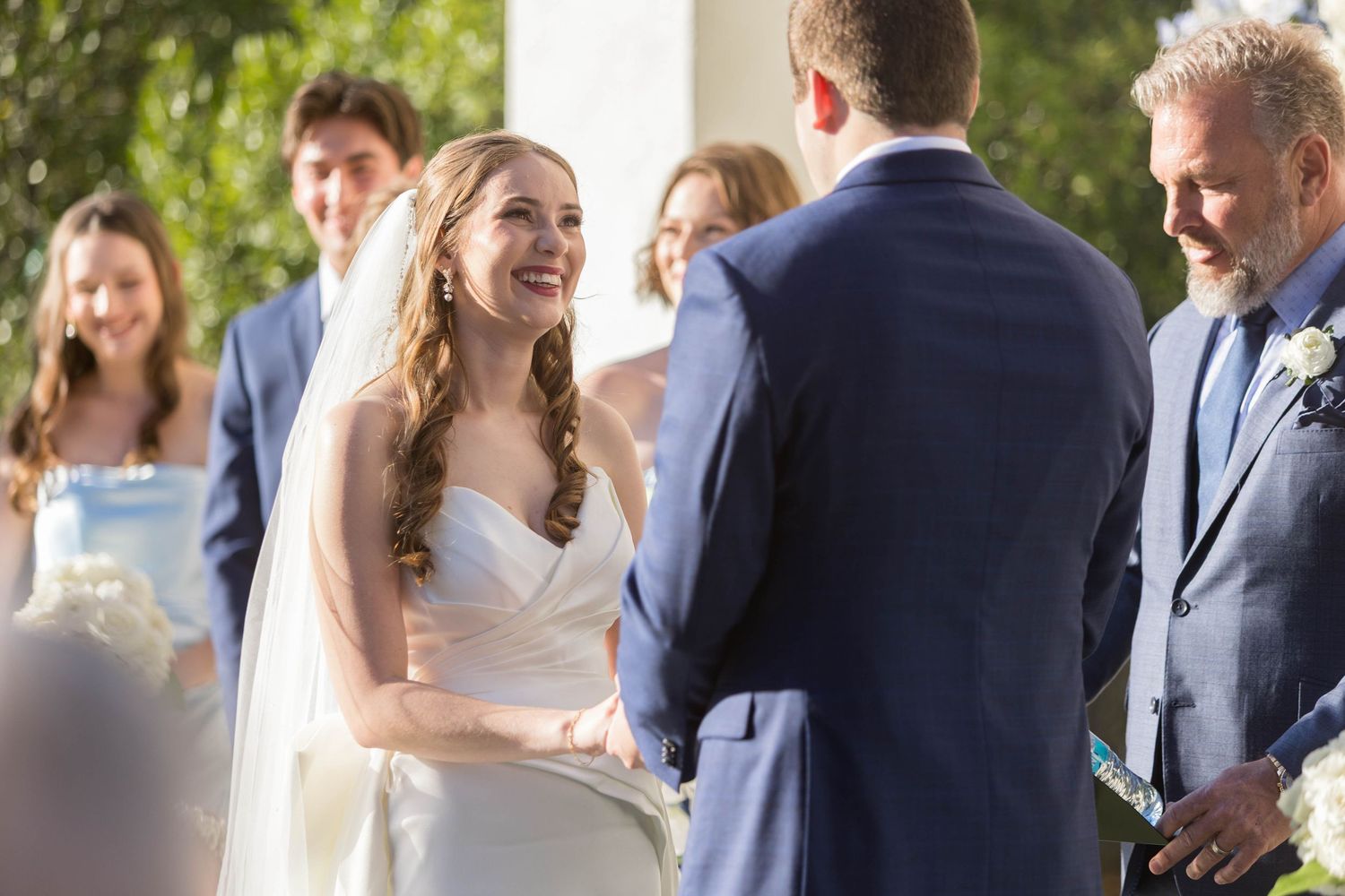 Bride smiling joyfully at groom during outdoor wedding ceremony with guests and officiant in background.