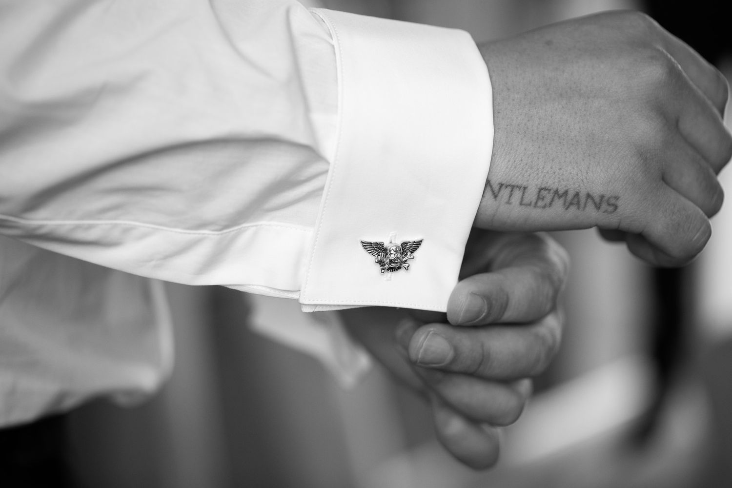 Black and white photo of a man's cufflink on a white dress shirt, with a GENTLEMANS tattoo visible on his wrist.