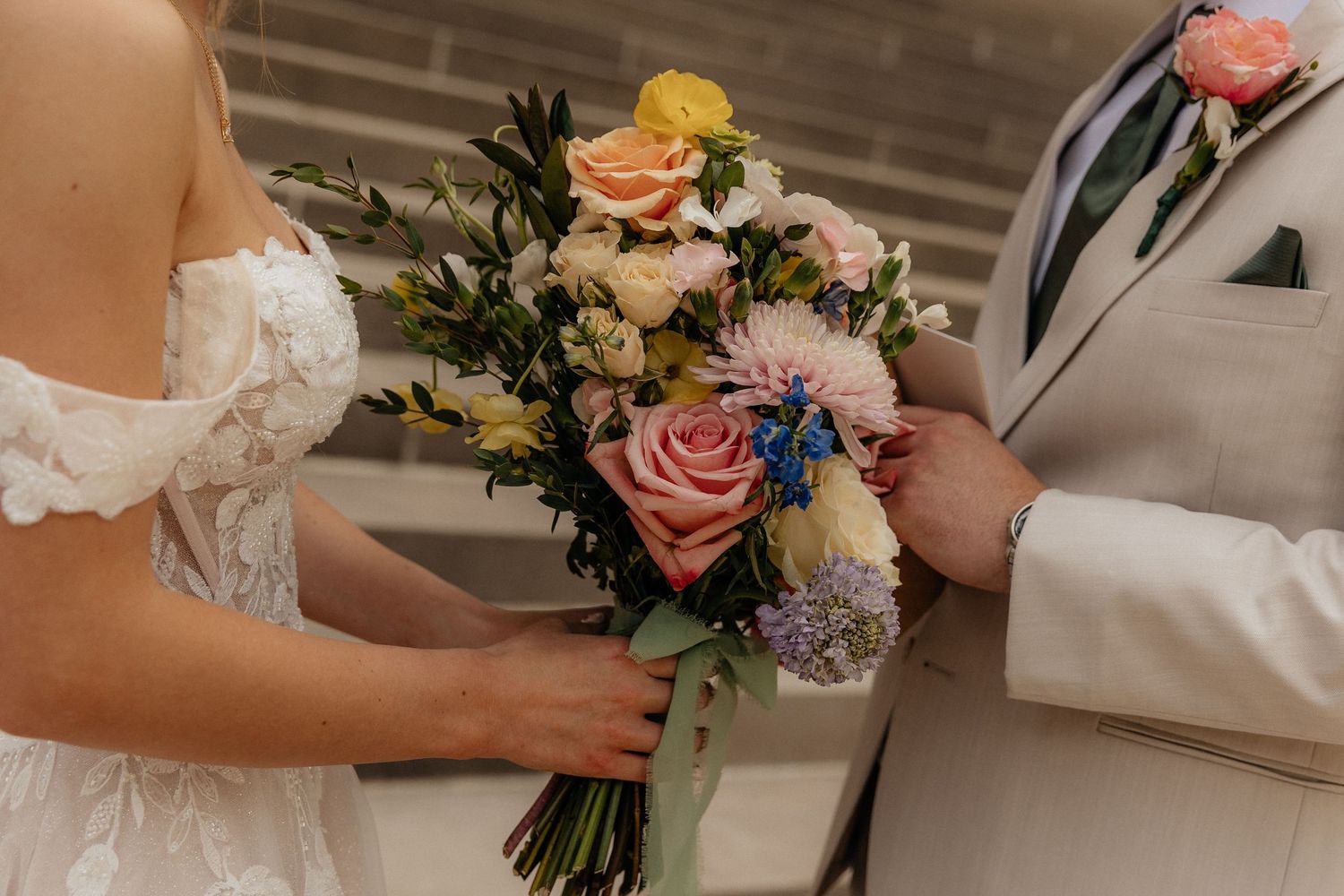Bride in white lace gown and groom in tan suit share a colorful wedding bouquet of pink, blue, and yellow flowers.