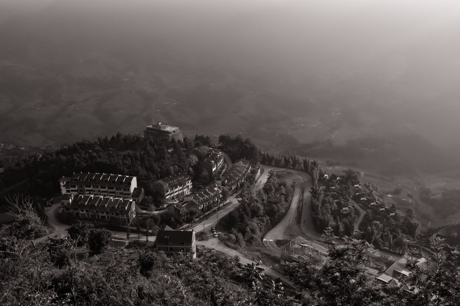 Aerial black and white view of a mountain resort with winding roads surrounded by misty valleys.
