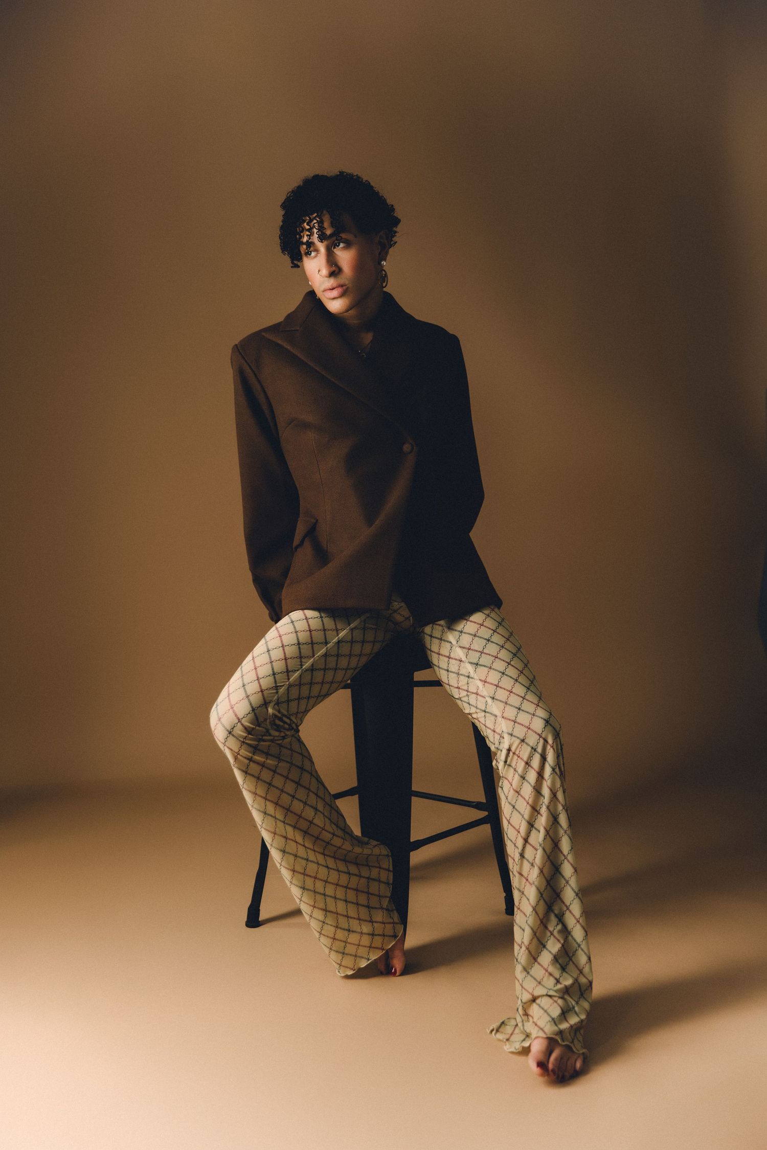 Model in brown blazer and plaid flare pants sits on stool against beige background in fashion editorial.