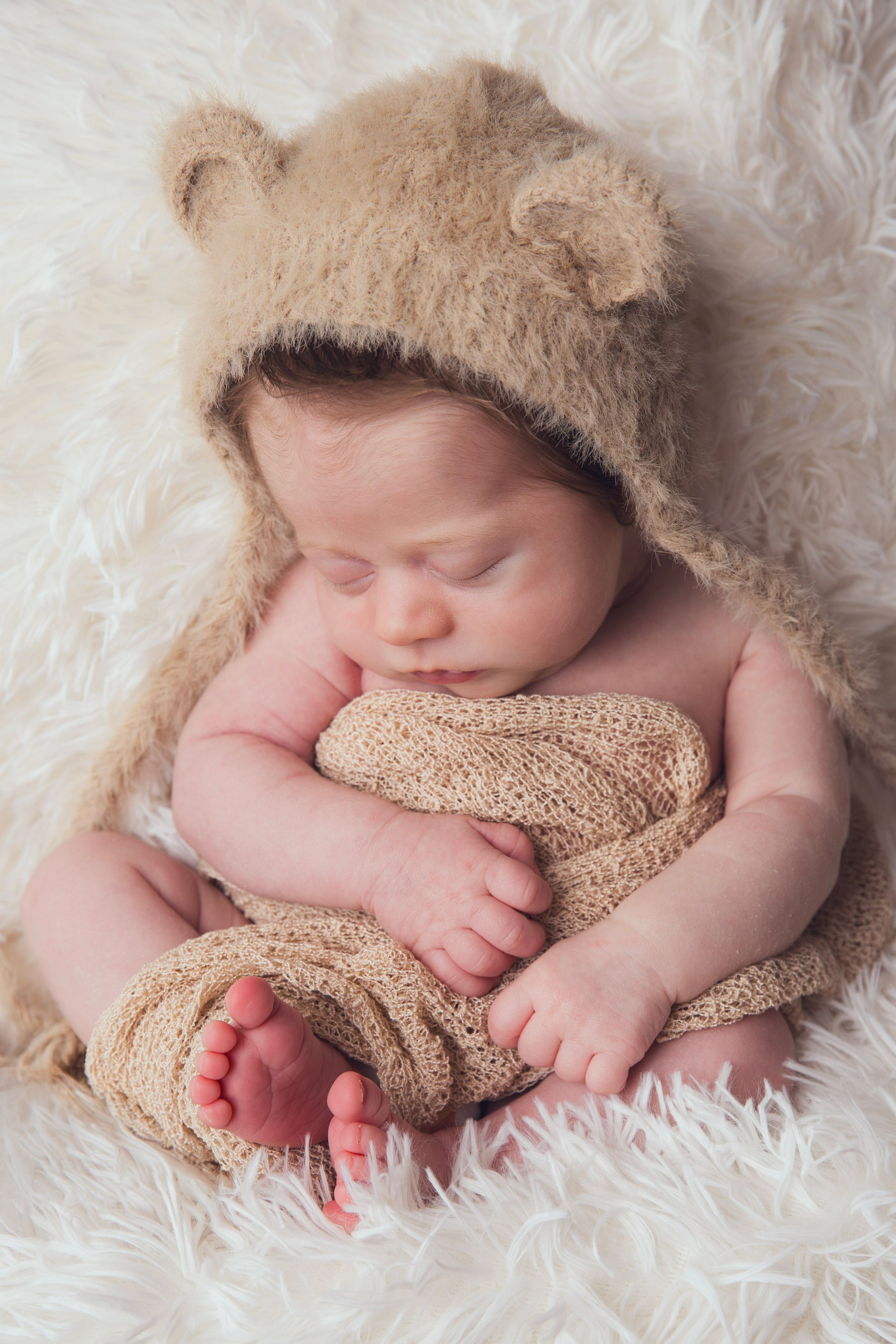 Sleeping newborn baby in a fuzzy bear costume with ears, resting on a soft white fur blanket.