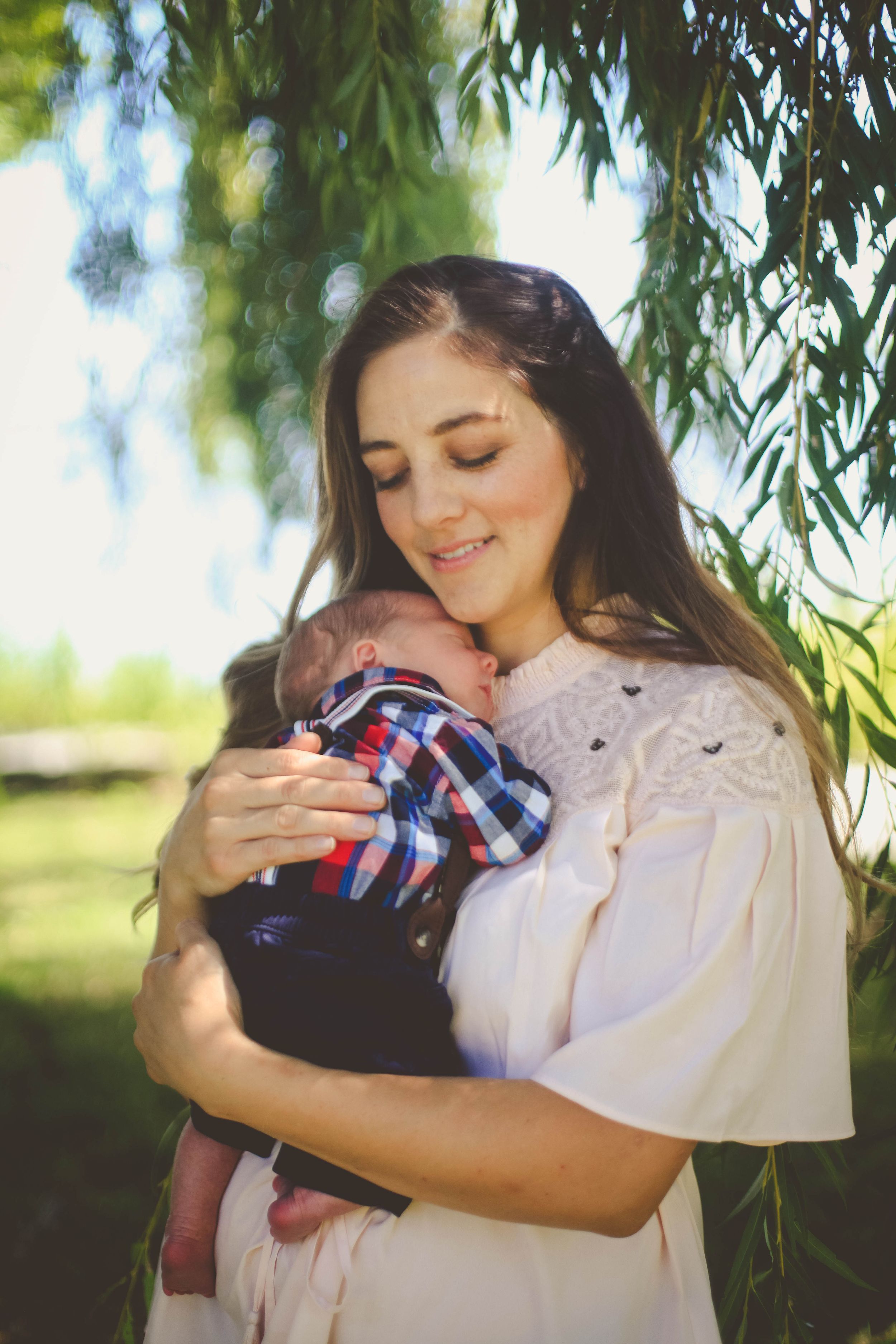 Young mother in white dress tenderly holds newborn baby outdoors under willow tree in warm sunlight.