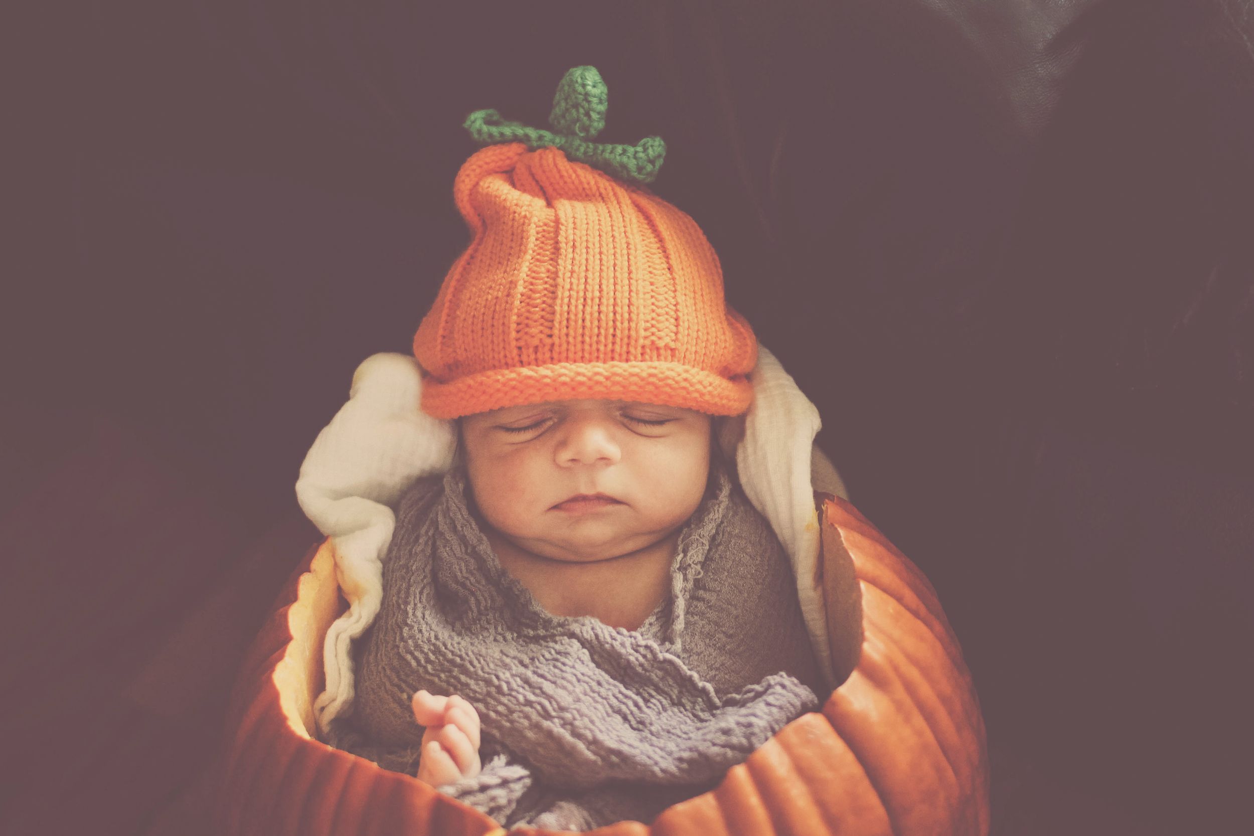 Newborn baby sleeping peacefully in an orange pumpkin costume with a knit pumpkin stem hat for Halloween.