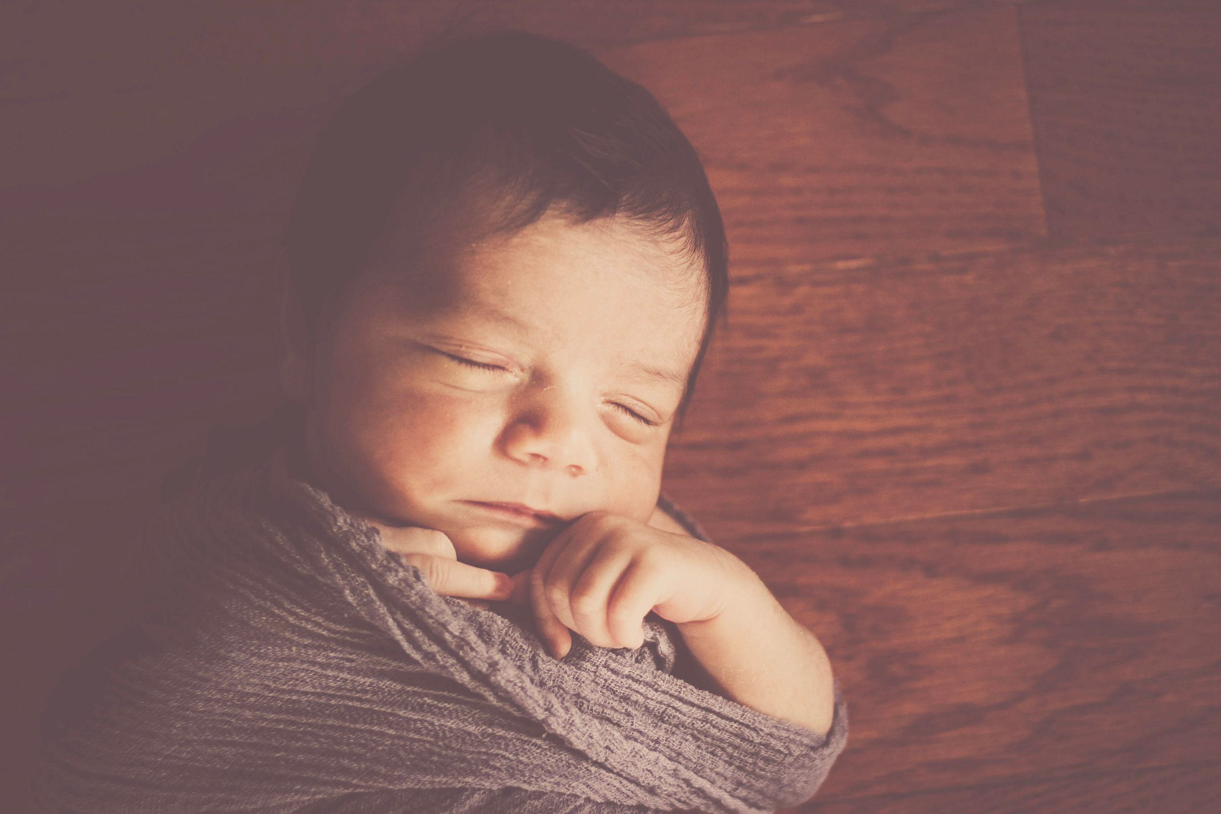 Peaceful newborn baby swaddled in gray wrap sleeping on rustic wooden surface in soft warm light.