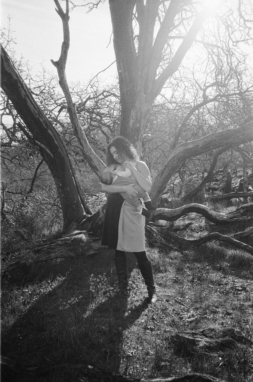 Woman holding baby stands in sunlit glade among bare twisted trees, black and white film photo.