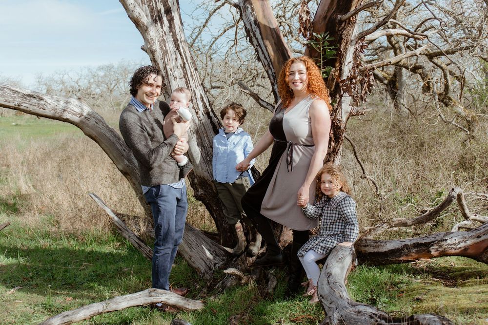 Family of four standing and sitting on a large gnarled tree trunk in a sunny meadow.