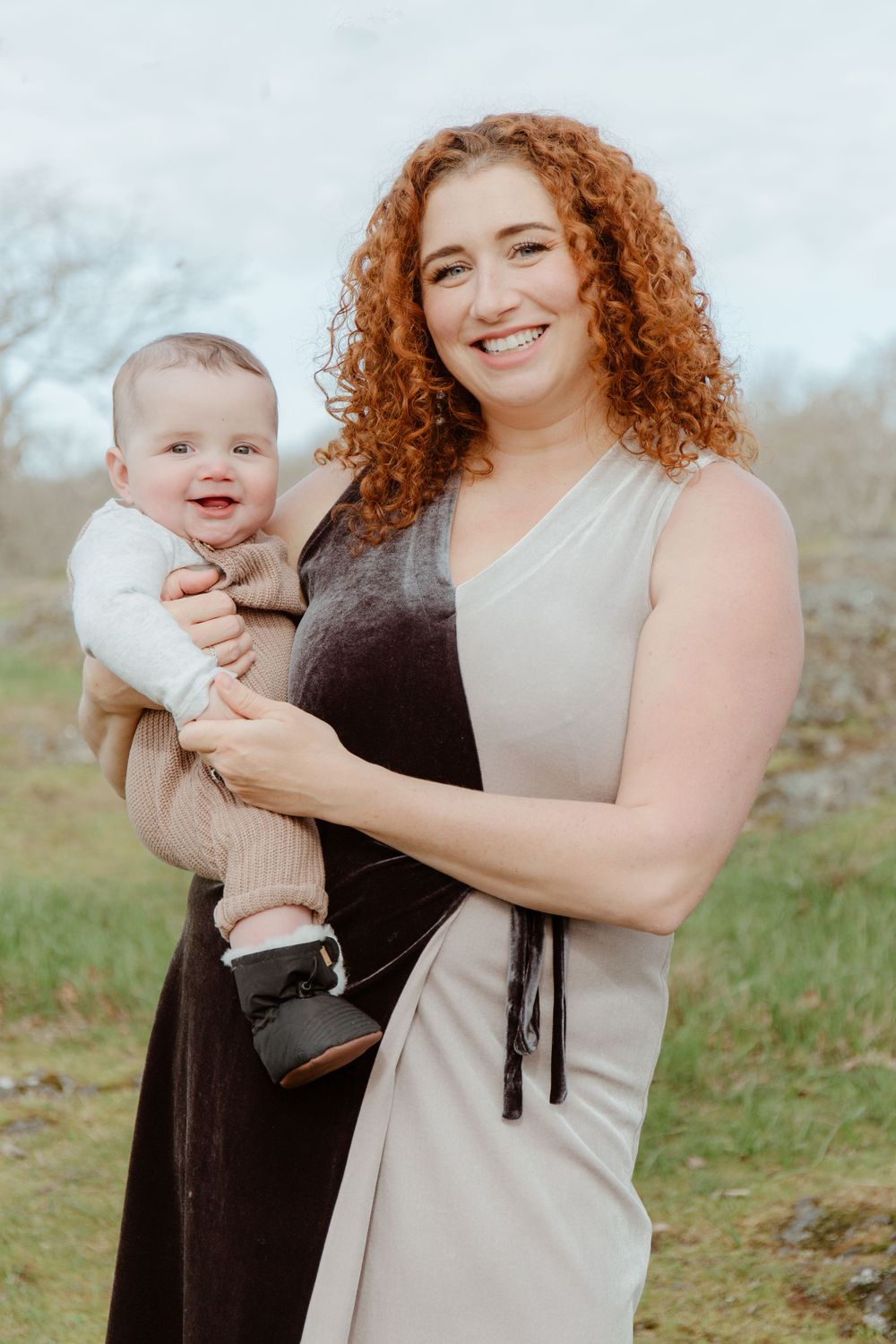 Smiling redhead woman with curly hair holding a happy baby outdoors in a green field.