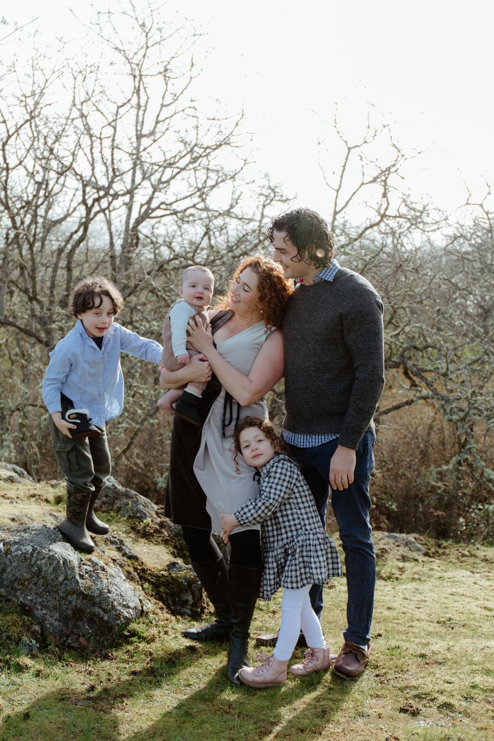 Joyful family of five sharing a candid moment outdoors among bare winter trees on a grassy hillside.