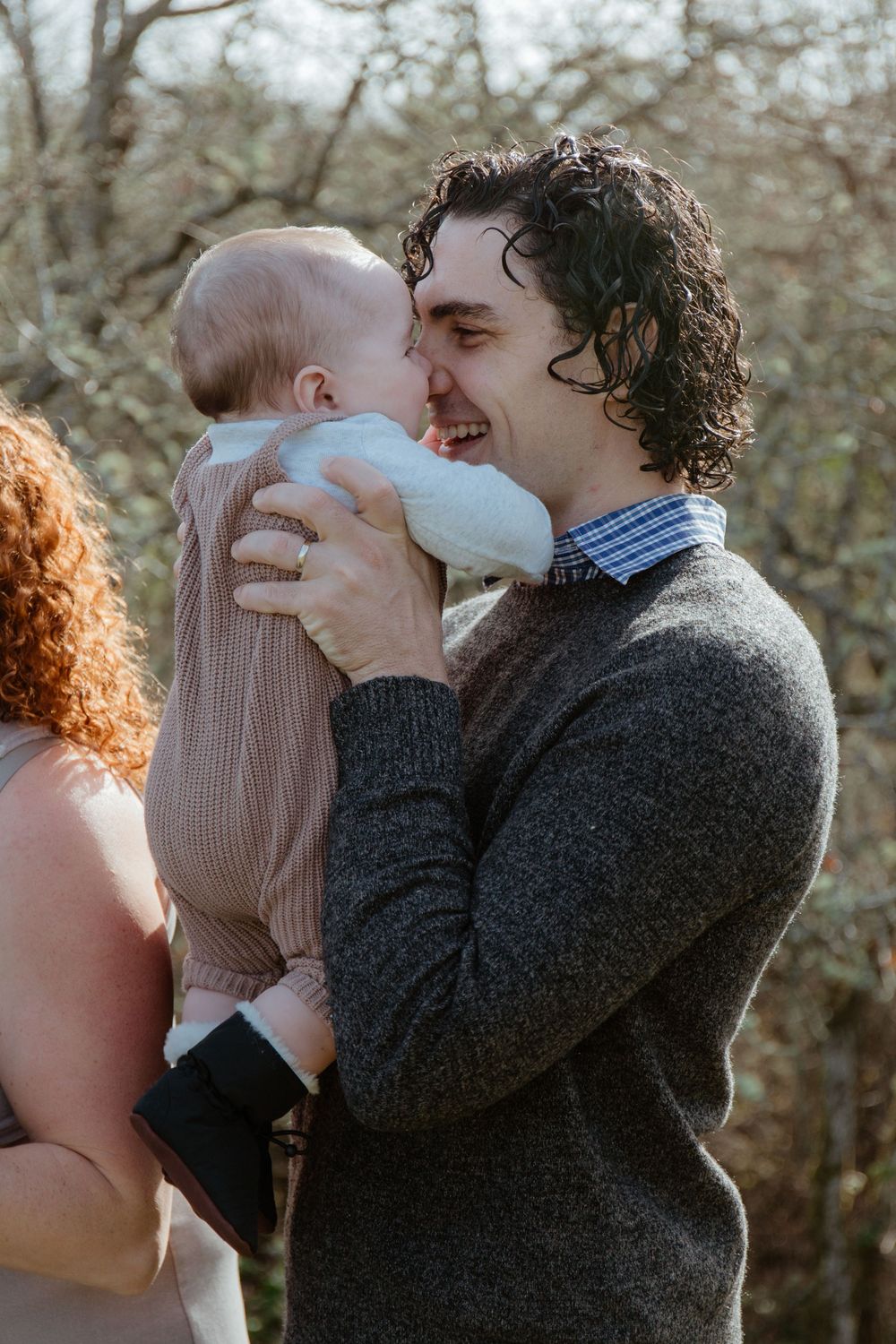 Smiling father in gray sweater lovingly holds baby up to his face outdoors in natural light.