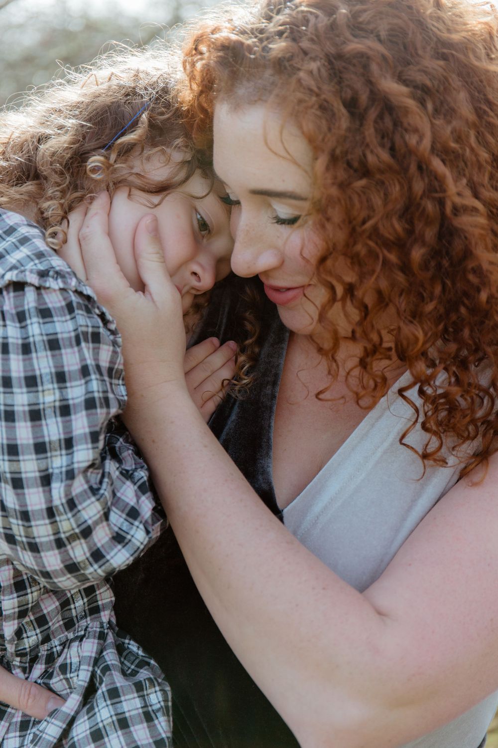 Tender moment between curly red-haired mother and young daughter embracing cheek to cheek outdoors.