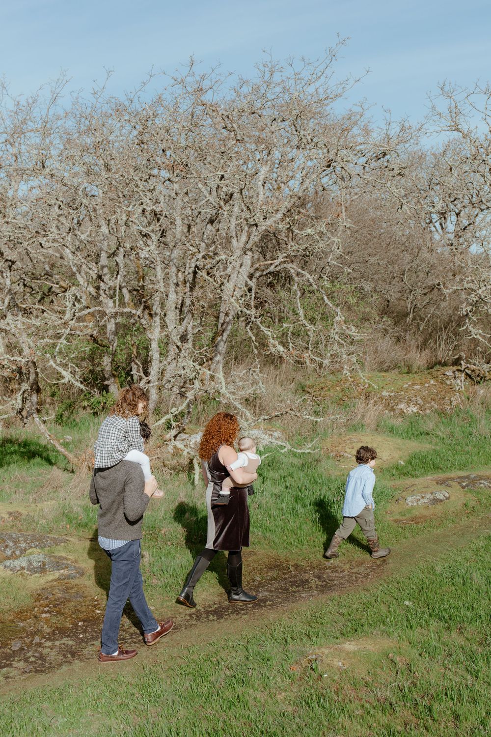 Family of five walking along a grassy path surrounded by bare trees on a sunny day.