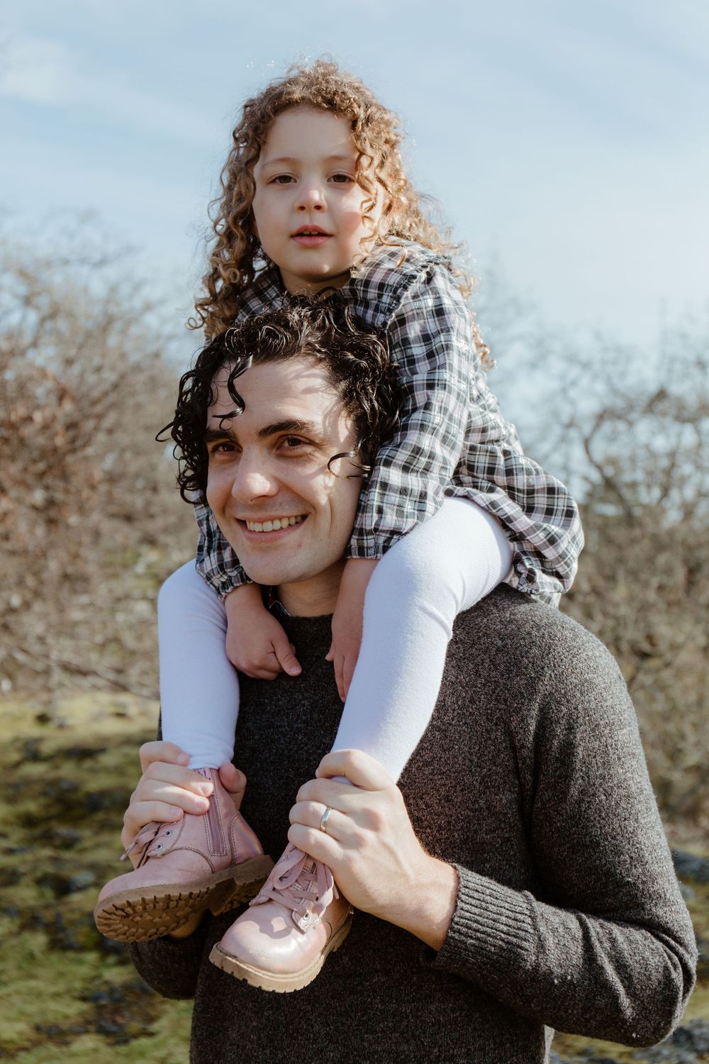 Smiling father carrying young curly-haired daughter on his shoulders outdoors against a blue sky.