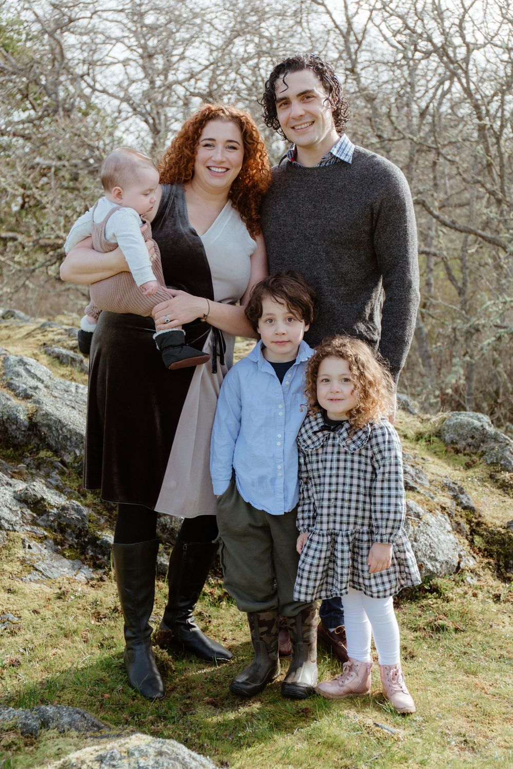 Family of five posing outdoors on mossy rocks, parents with three young children smiling in casual attire.