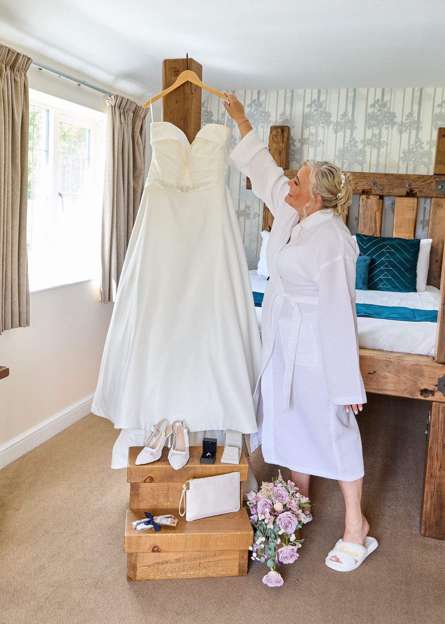 Bride in white robe admiring her strapless wedding gown hanging in a decorated bedroom with accessories displayed below.