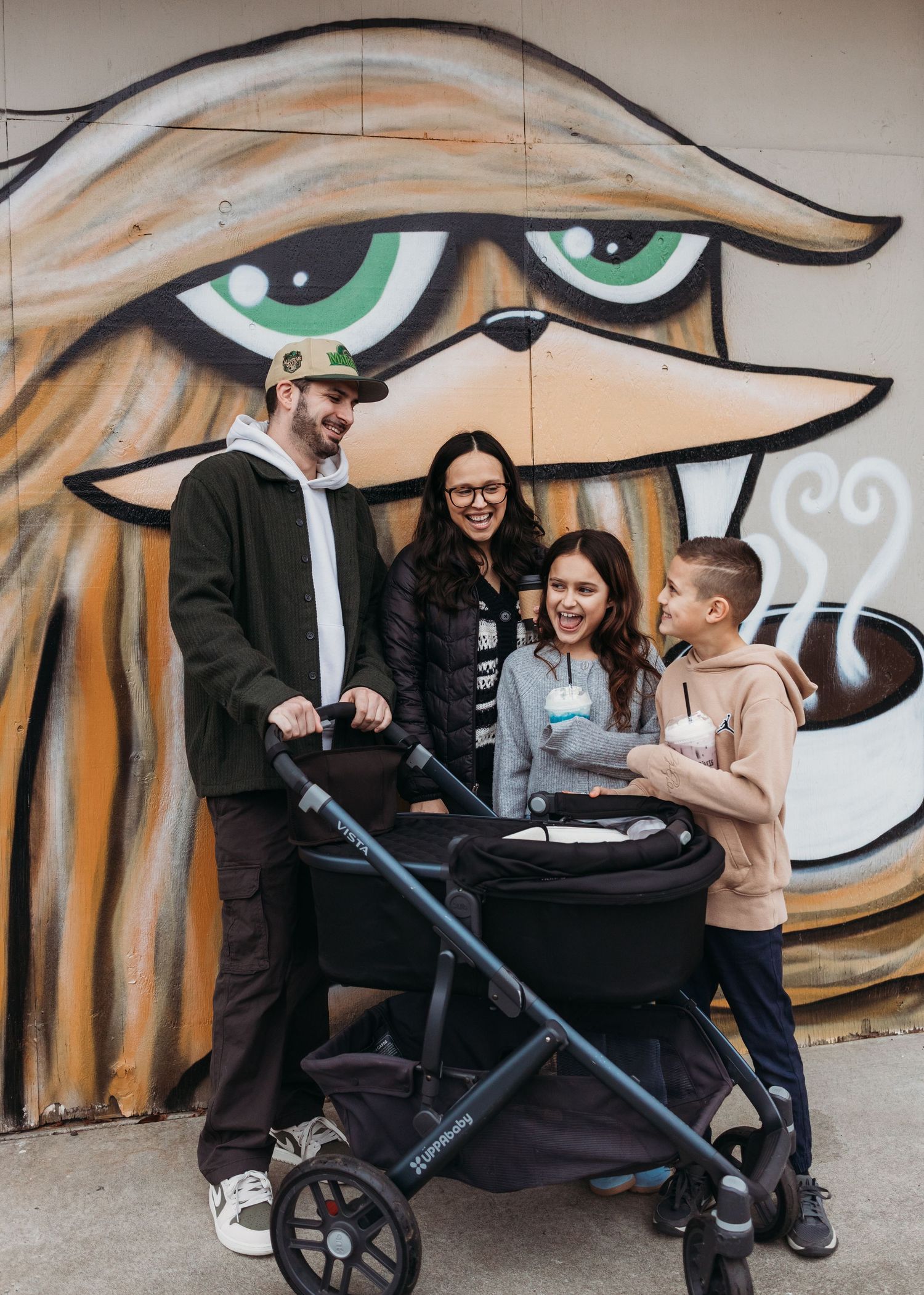 Family of four with stroller laughing in front of colorful street art mural outdoors.