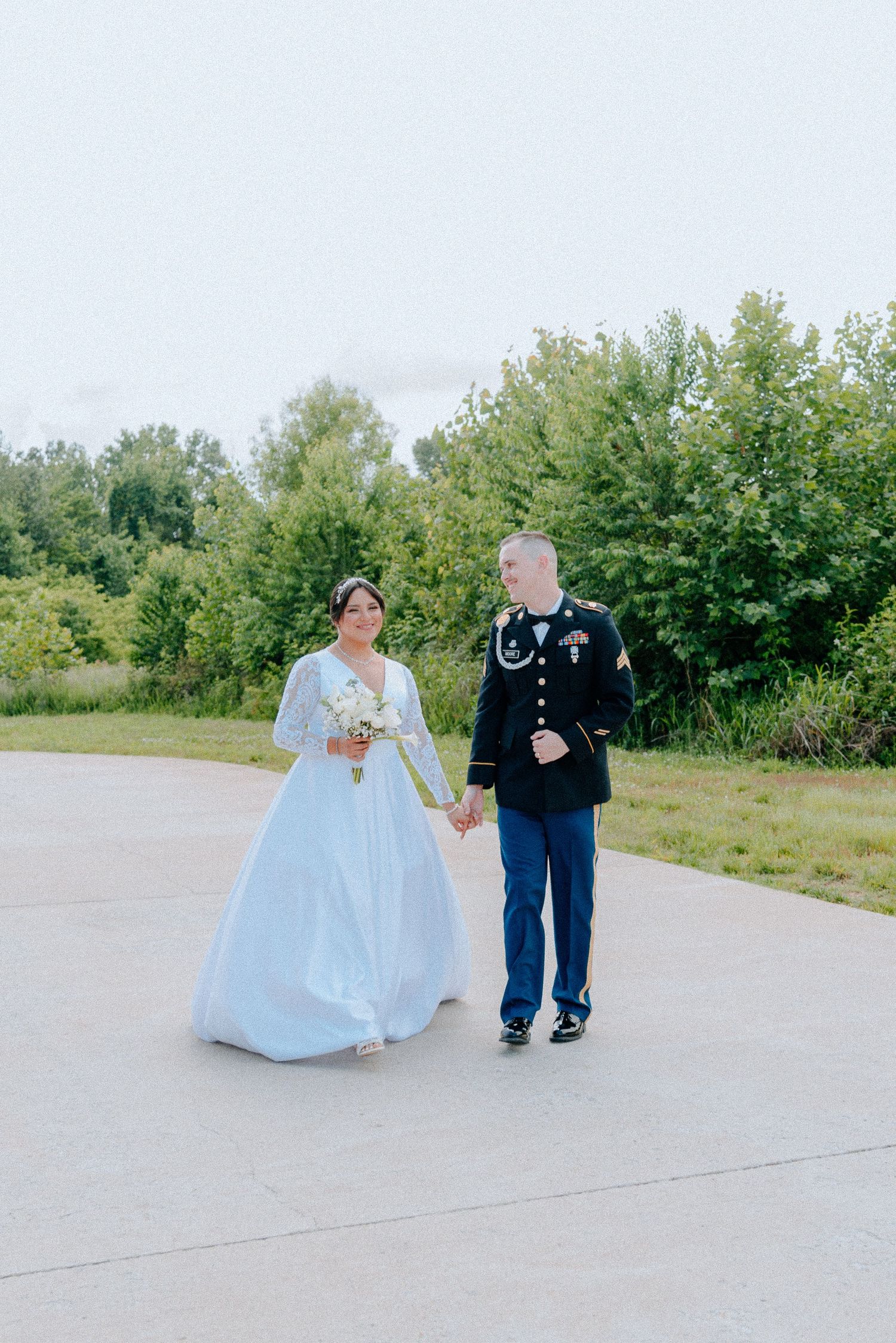 Military groom in dress uniform twirls bride in white lace wedding gown outdoors on sunny day.
