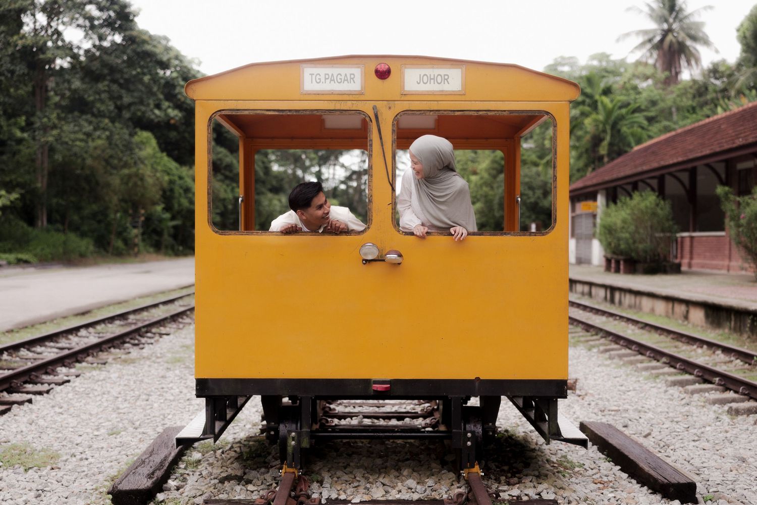 Two people lean out of a yellow vintage train car on railway tracks surrounded by lush greenery.