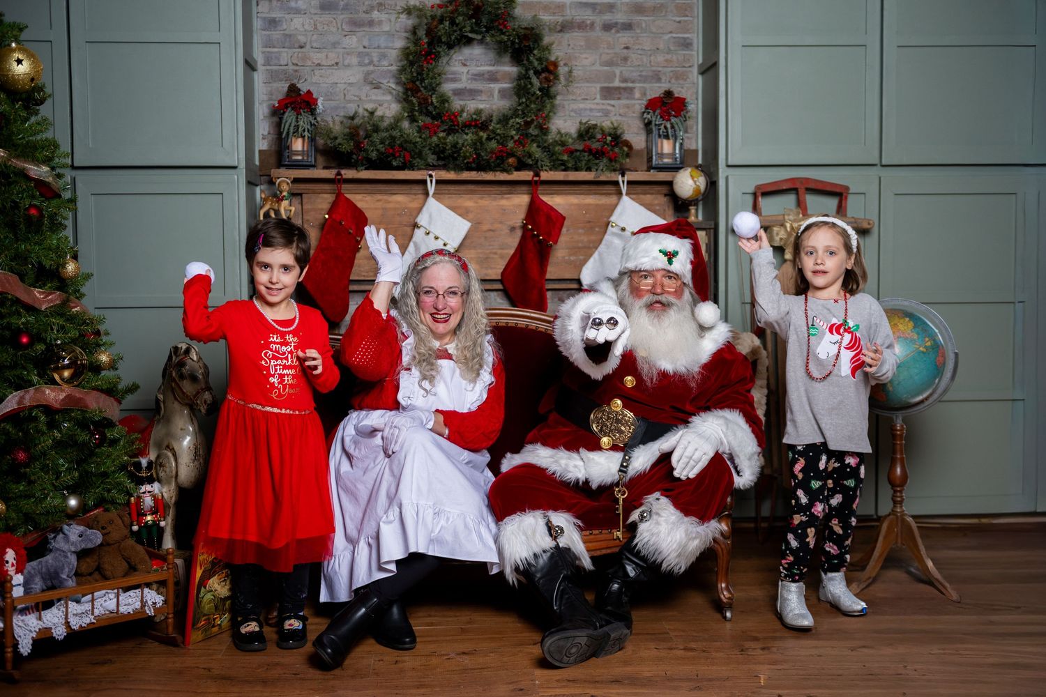 Two children pose with Santa Claus and Mrs. Claus by a decorated fireplace and Christmas tree in a festive holiday photo.