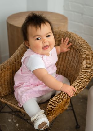 Smiling baby in pink seersucker romper sitting in wicker chair, wearing white shoes, looking up happily.