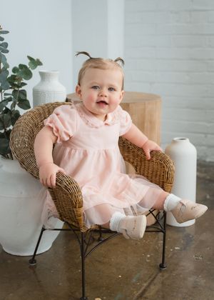 Baby girl in pink dress with tulle skirt sits in wicker chair, wearing white socks and mary janes.