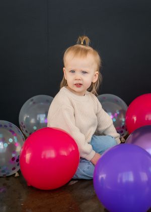 Toddler with blonde hair sits among colorful pink, purple, and clear confetti balloons against a dark background.