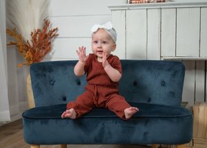 Adorable baby girl in rust romper and white bow headband sitting on a teal velvet mini sofa.