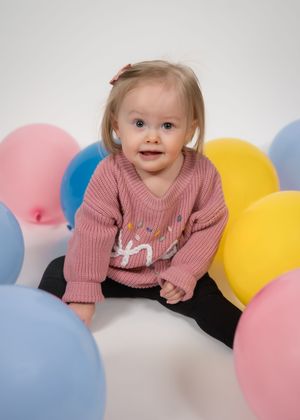 Smiling toddler girl in pink sweater sitting among colorful balloons on white background.