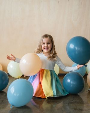 Happy young girl in rainbow dress surrounded by colorful balloons, laughing and playing in a bright studio setting.
