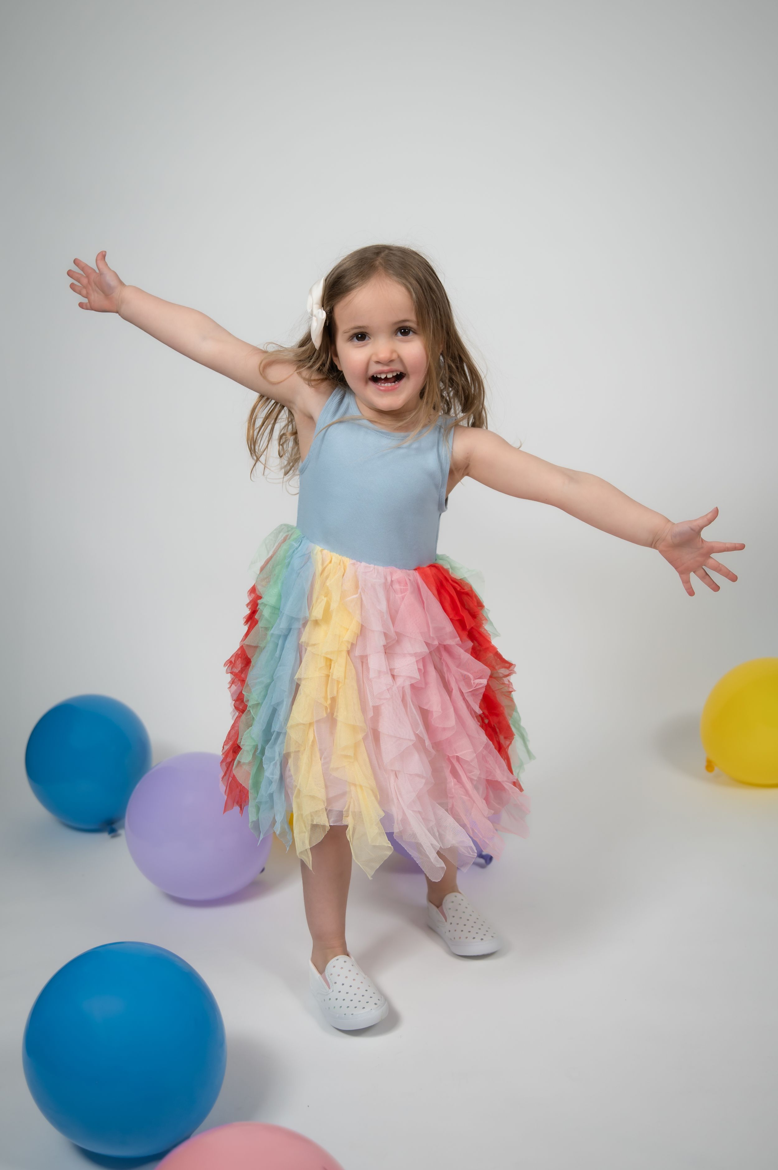 Happy little girl in colorful rainbow tutu dress with arms outstretched, surrounded by balloons on white background.