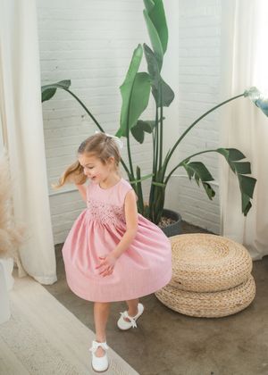 A young girl twirls in a pink smocked dress with white Mary Janes, posing near a tropical plant indoors.