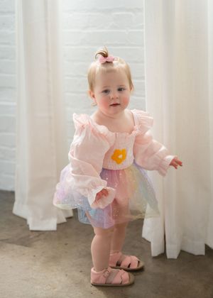 Baby girl in pink ruffled top, rainbow tutu skirt, and sandals with flower applique, standing by white curtains.